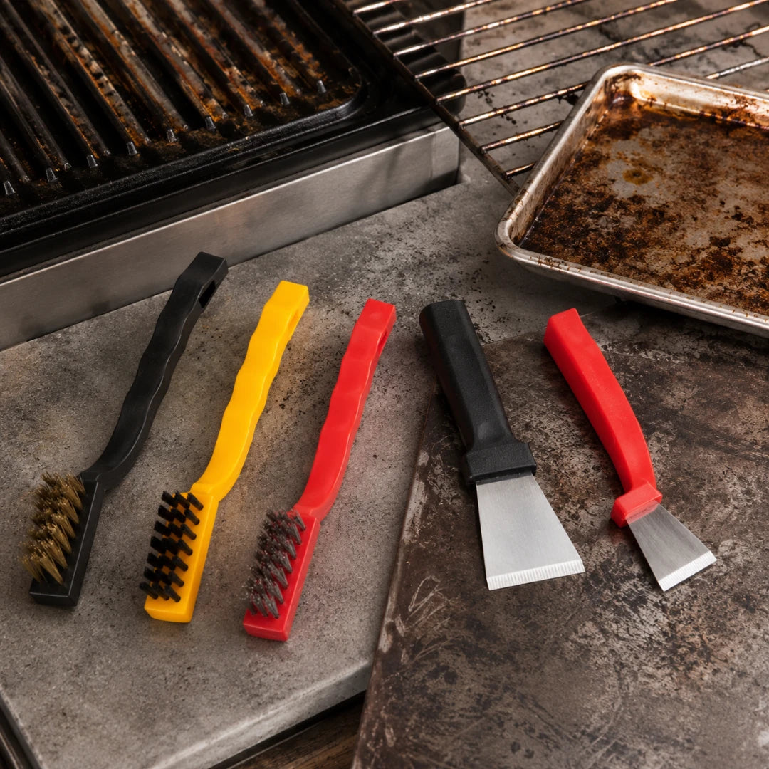 Josh & Tate - ScrubEdge™ 5-Piece Cleaning Set - Four barbecue cleaning tools arranged on a stone surface beside a grill: two wire brushes in black and yellow with dark bristles, and two scraper tools in black and red with silver metal blades. A dirty baking pan and grill grate are visible in the background.