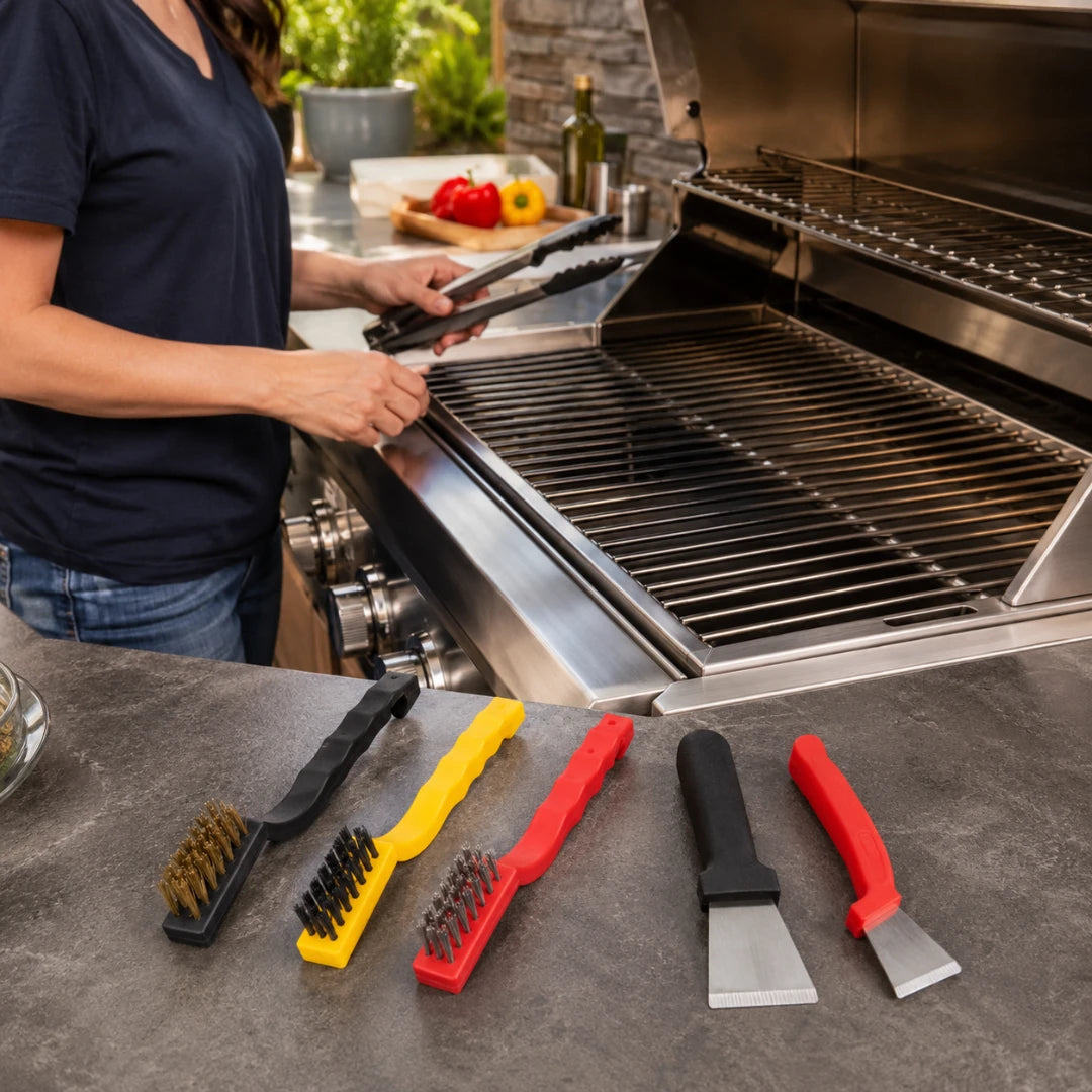 Josh & Tate -ScrubEdge™ 5-Piece Cleaning Set - Four grill cleaning tools are displayed on a stone countertop in front of an open stainless steel grill, with a person in a navy shirt using tongs to tend the grill in the background.