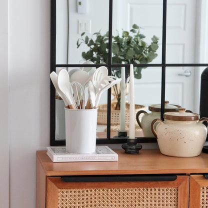 White utensil holder with kitchen utensils on a wooden cabinet with a mirror and plant in the background