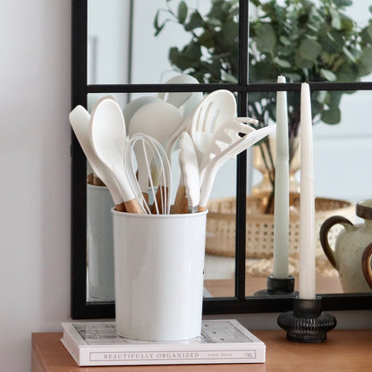 White utensil holder with kitchen utensils on a table next to candles and a book.