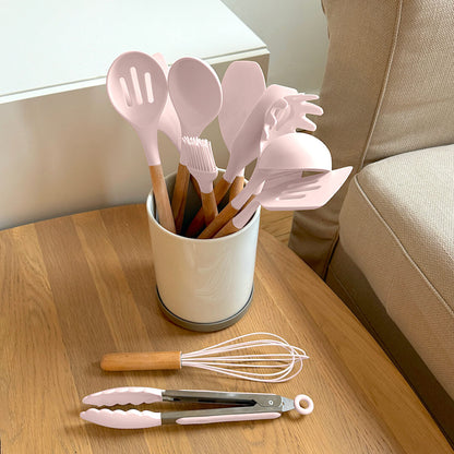 Set of pink kitchen utensils including spatulas, tongs, and a whisk on a wooden surface.