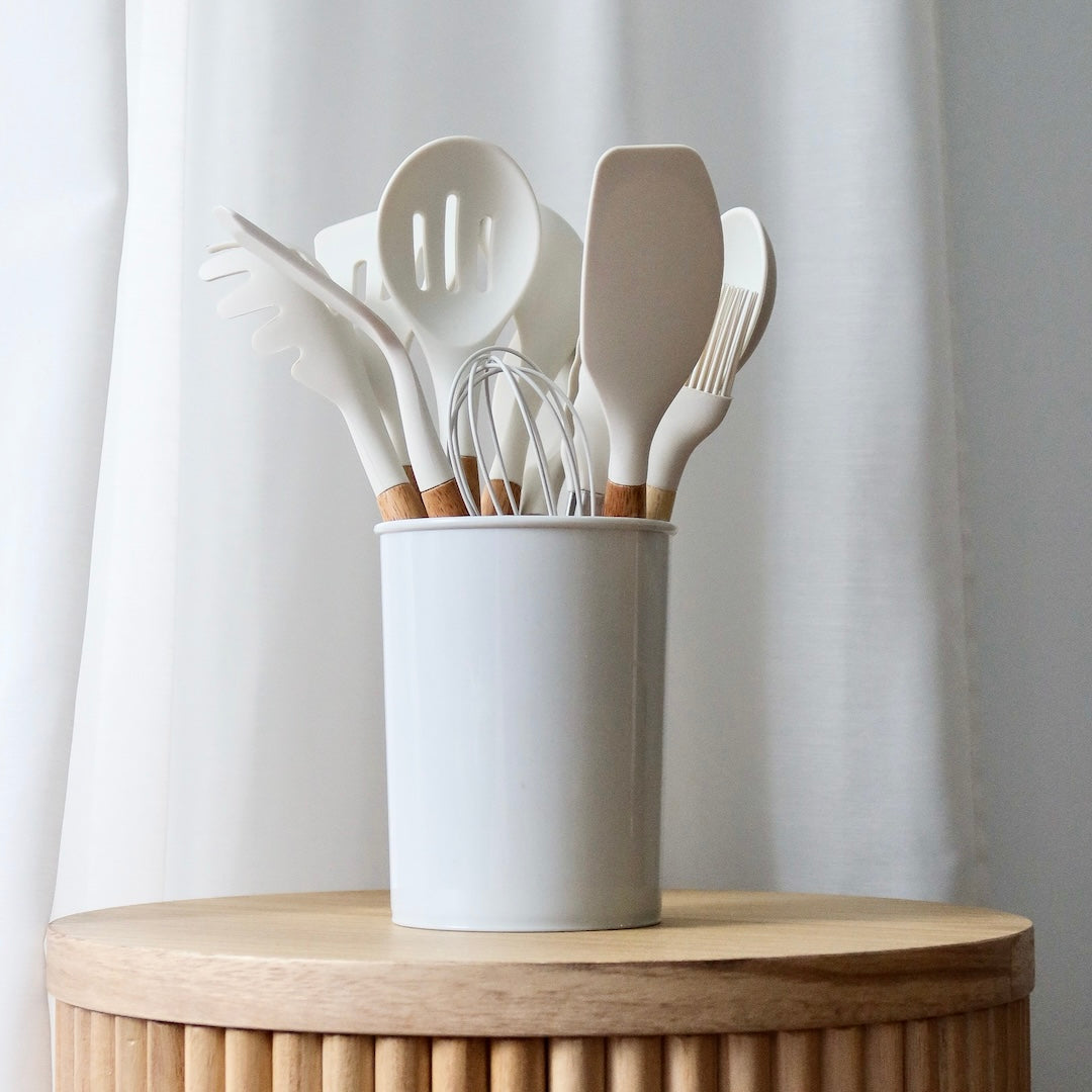 Set of kitchen utensils in a white container on a wooden surface with a light curtain background