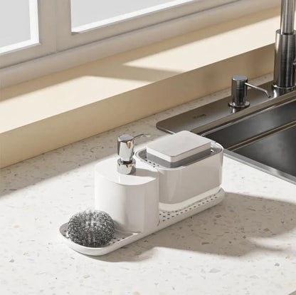 White kitchen sink caddy with soap dispenser, sponge holder, and scrubber on a light-colored countertop.