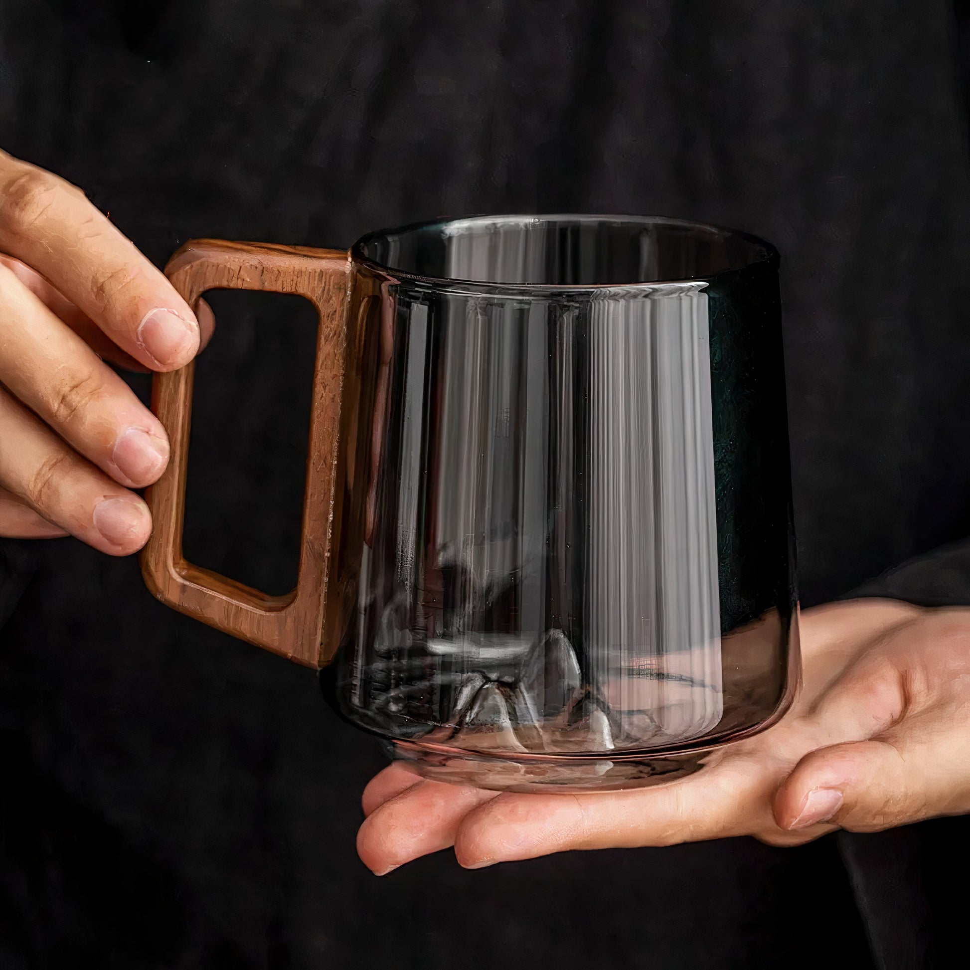 Clear glass mug with wooden handle held by hands against a dark background
