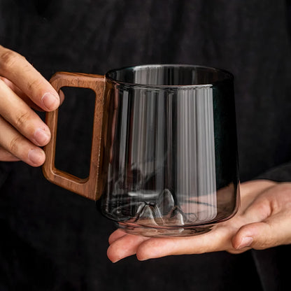 Clear glass mug with wooden handle held by hands against a dark background