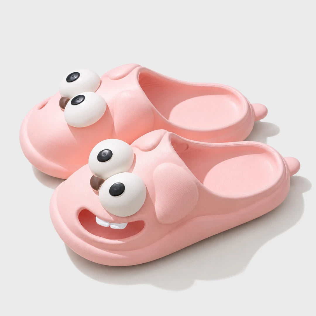 Josh & Tate - SnaggleToe™ Slides - A pair of pink slip-on slippers shaped like cartoon animal faces, with large white eyes and a smiling mouth on the uppers, photographed on a clean light gray background.