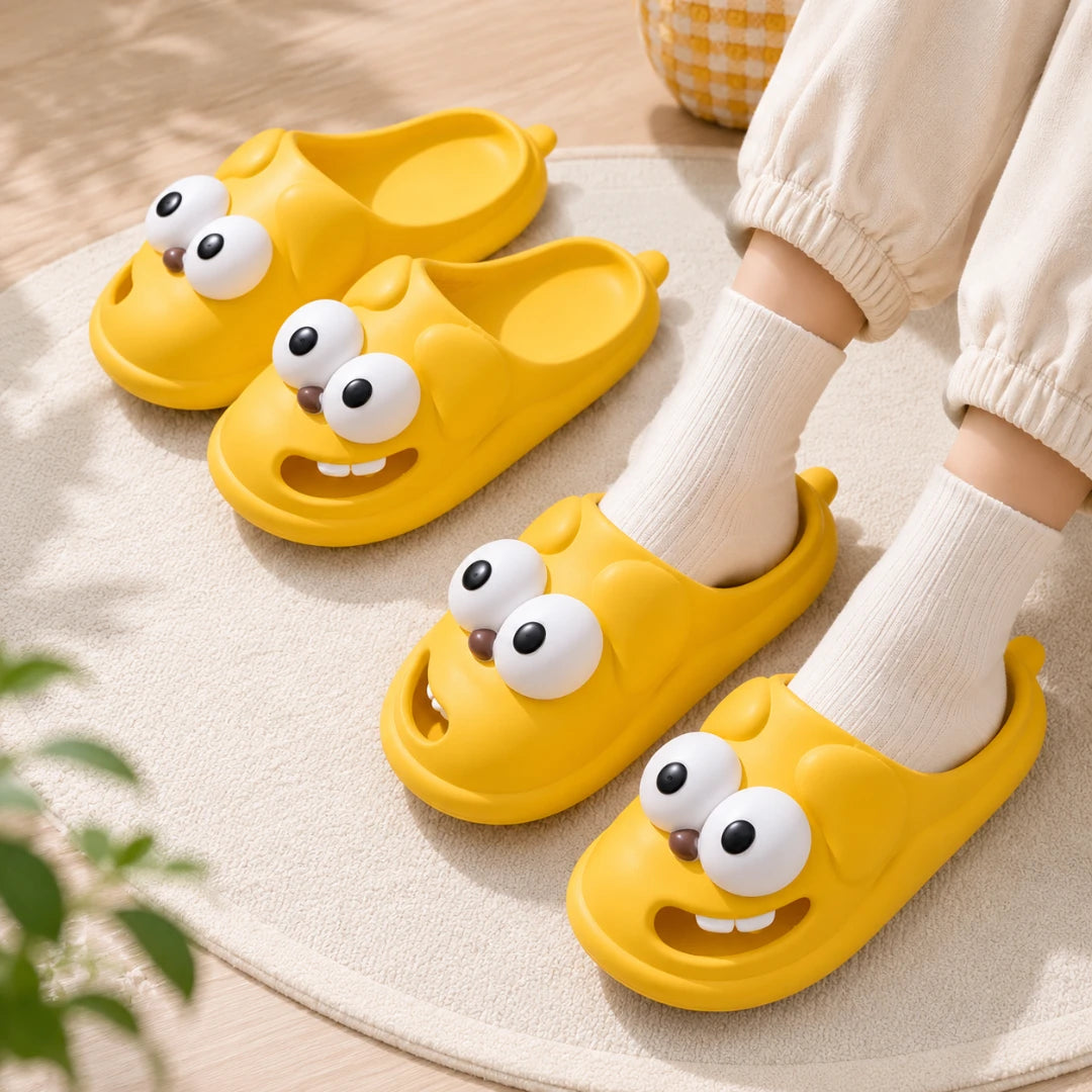 Josh & Tate - SnaggleToe™ Slides - Three bright yellow cartoon slip-on slippers with large white eyes and smiling mouths are shown on a light rug. Two slippers are displayed upright, and one is being worn with white socks and cream pants visible.