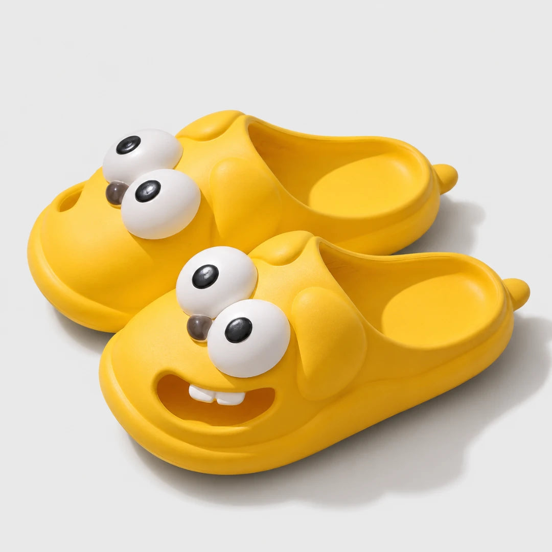 Josh & Tate - SnaggleToe™ Slides - A pair of bright yellow children's slip-on slippers shaped like cartoon animal heads with large white eyes and small black pupils, shown on a light gray background.