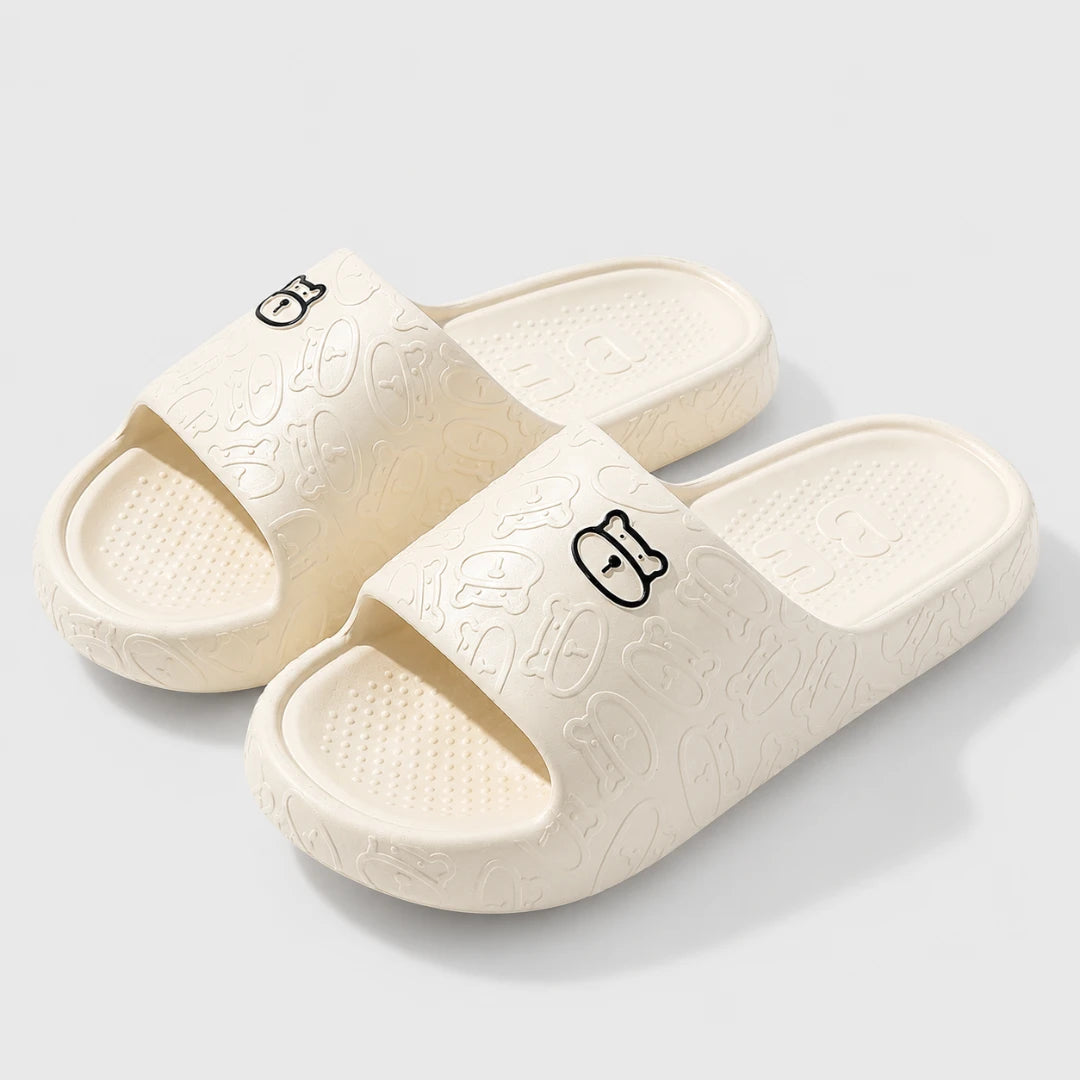 Josh & Tate - SoftBear™ Non-Slip Cushion Slides - A pair of off-white slide sandals with embossed cartoon patterns and small black cartoon face logos on the straps, shown on a light gray background.