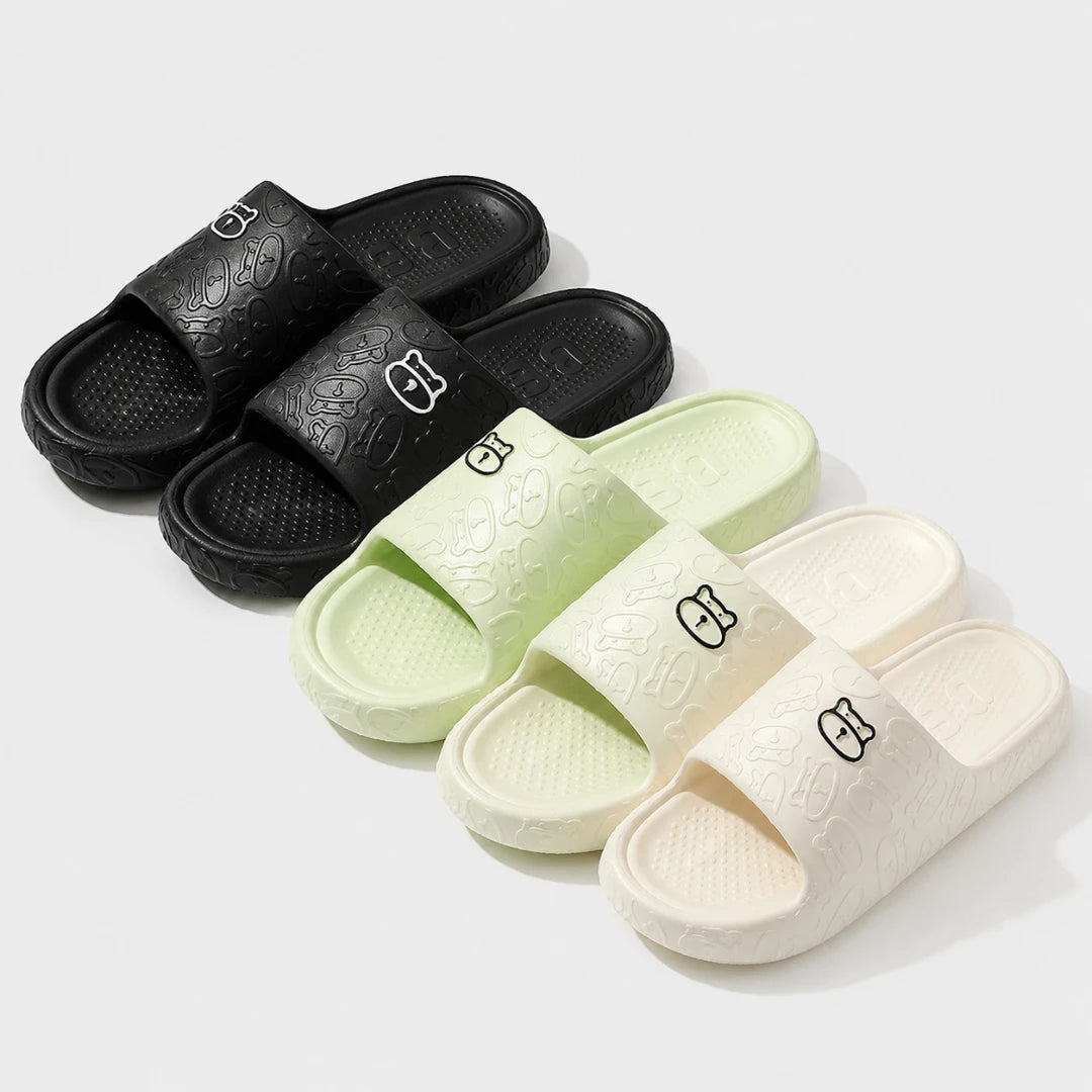 Josh & Tate - SoftBear™ Non-Slip Cushion Slides - Three pairs of slide sandals arranged on a light gray background: black, pale mint green, and off-white. Each sandal has an embossed textured pattern and a small stylized logo on the strap.