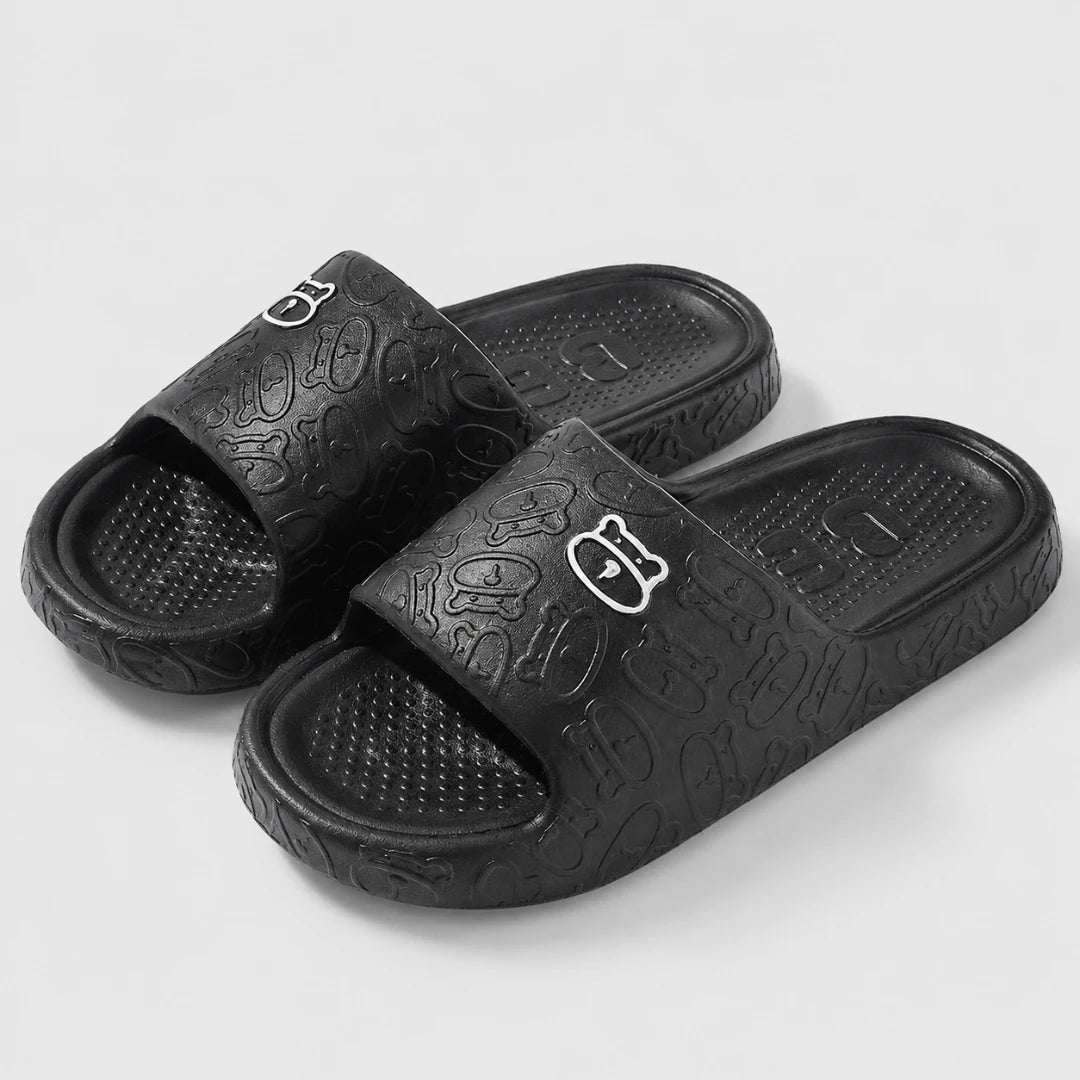 Josh & Tate - SoftBear™ Non-Slip Cushion Slides - A pair of black slide sandals with an embossed abstract pattern and a small white stylized logo on the strap, shown on a light gray background.