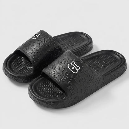 Josh & Tate - SoftBear™ Non-Slip Cushion Slides - A pair of black slide sandals with an embossed abstract pattern and a small white stylized logo on the strap, shown on a light gray background.