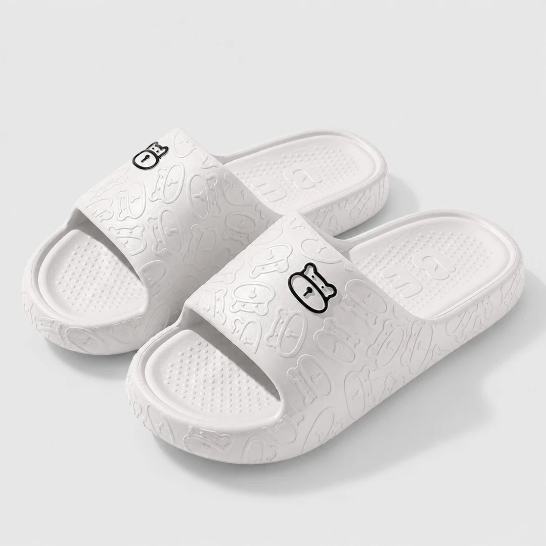 Josh & Tate - SoftBear™ Non-Slip Cushion Slides - A pair of white slide sandals with a textured, embossed cartoon pattern and a small black outline character logo on the strap, shown on a light gray background.