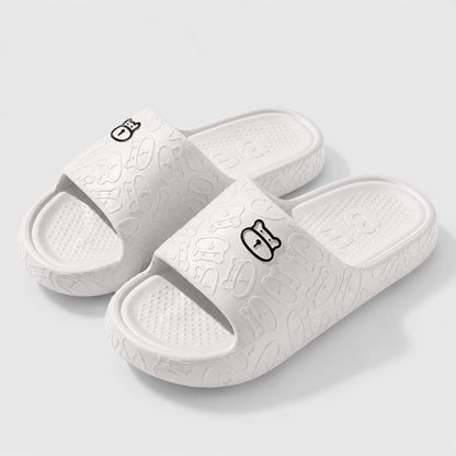 Josh & Tate - SoftBear™ Non-Slip Cushion Slides - A pair of white slide sandals with a textured, embossed cartoon pattern and a small black outline character logo on the strap, shown on a light gray background.