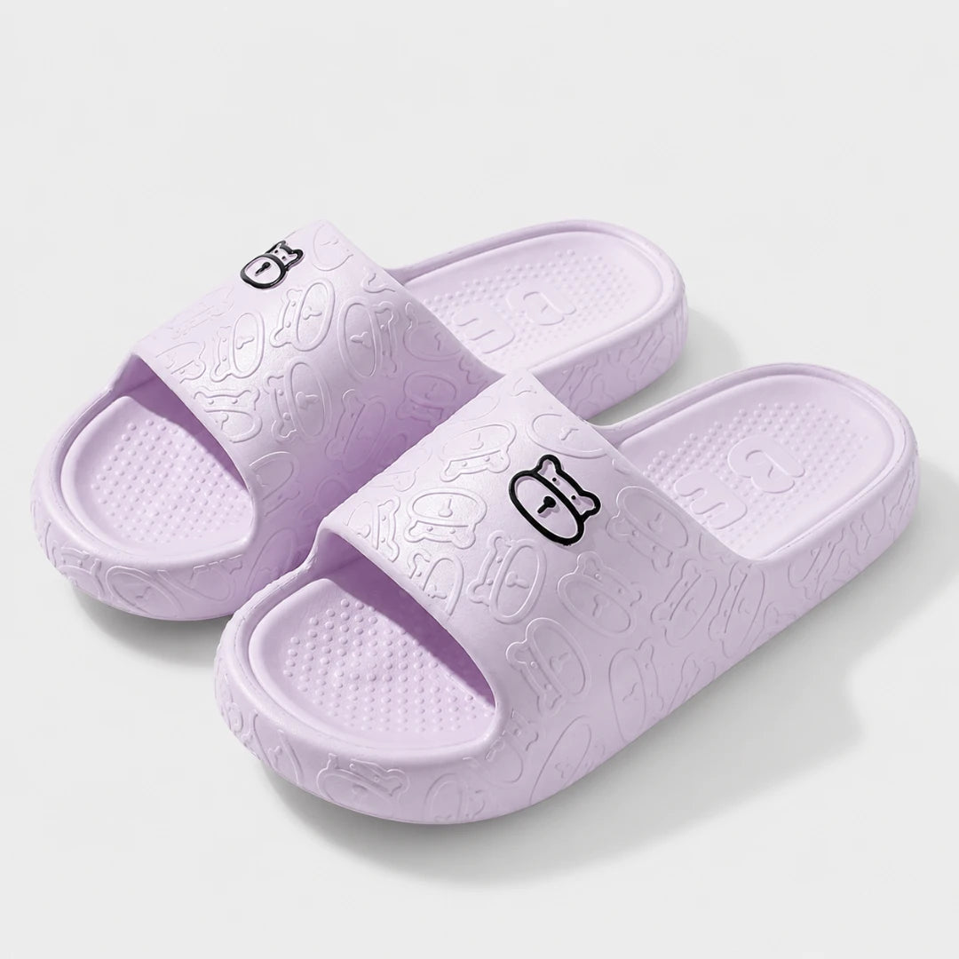 Josh & Tate - SoftBear™ Non-Slip Cushion Slides - A pair of light purple slide sandals with embossed cartoon patterns and small black outline character logos on the straps, shown on a light gray background.