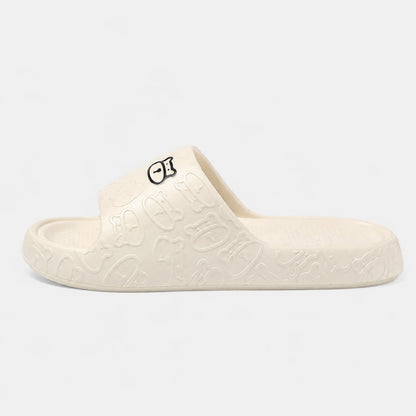 Josh & Tate - SoftBear™ Non-Slip Cushion Slides - A single cream-colored slide sandal with an embossed graffiti pattern and a small black and white graphic logo on the strap, shown against a plain light gray background.
