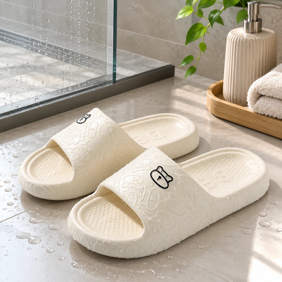 Josh & Tate - SoftBear™ Non-Slip Cushion Slides - A pair of off-white foam slide sandals with embossed floral patterns and small cartoon face details, displayed on a tiled bathroom floor with water droplets, a glass shower door, and a beige pump bottle in the background.