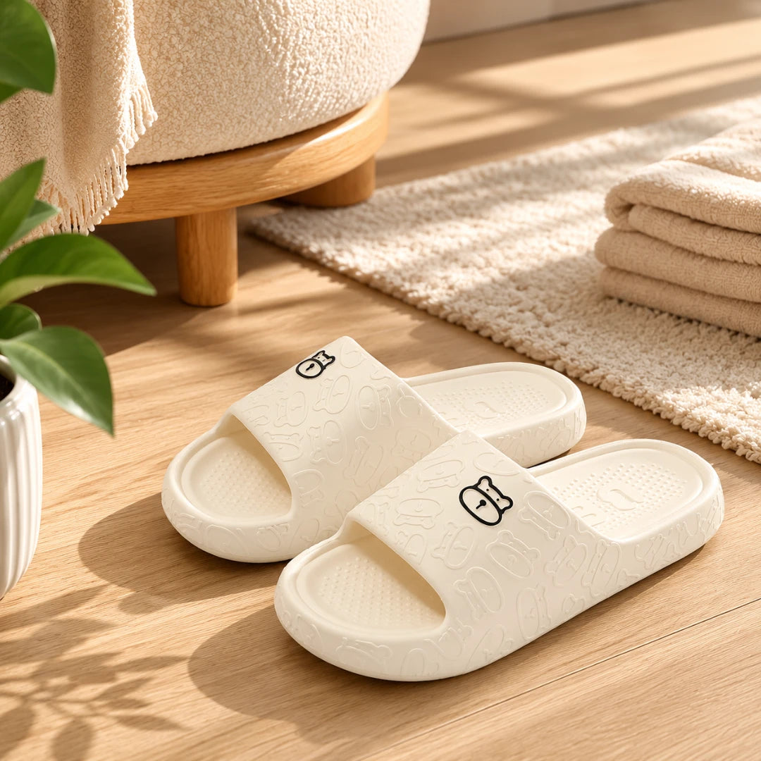 Josh & Tate - SoftBear™ Non-Slip Cushion Slides - A pair of off-white slide slippers with a textured pattern and small cartoon bunny details, displayed on a light wood floor in a cozy, sunlit room setting.
