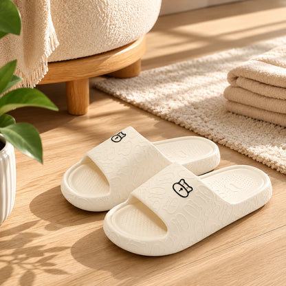 Josh & Tate - SoftBear™ Non-Slip Cushion Slides - A pair of off-white slide slippers with a textured pattern and small cartoon bunny details, displayed on a light wood floor in a cozy, sunlit room setting.