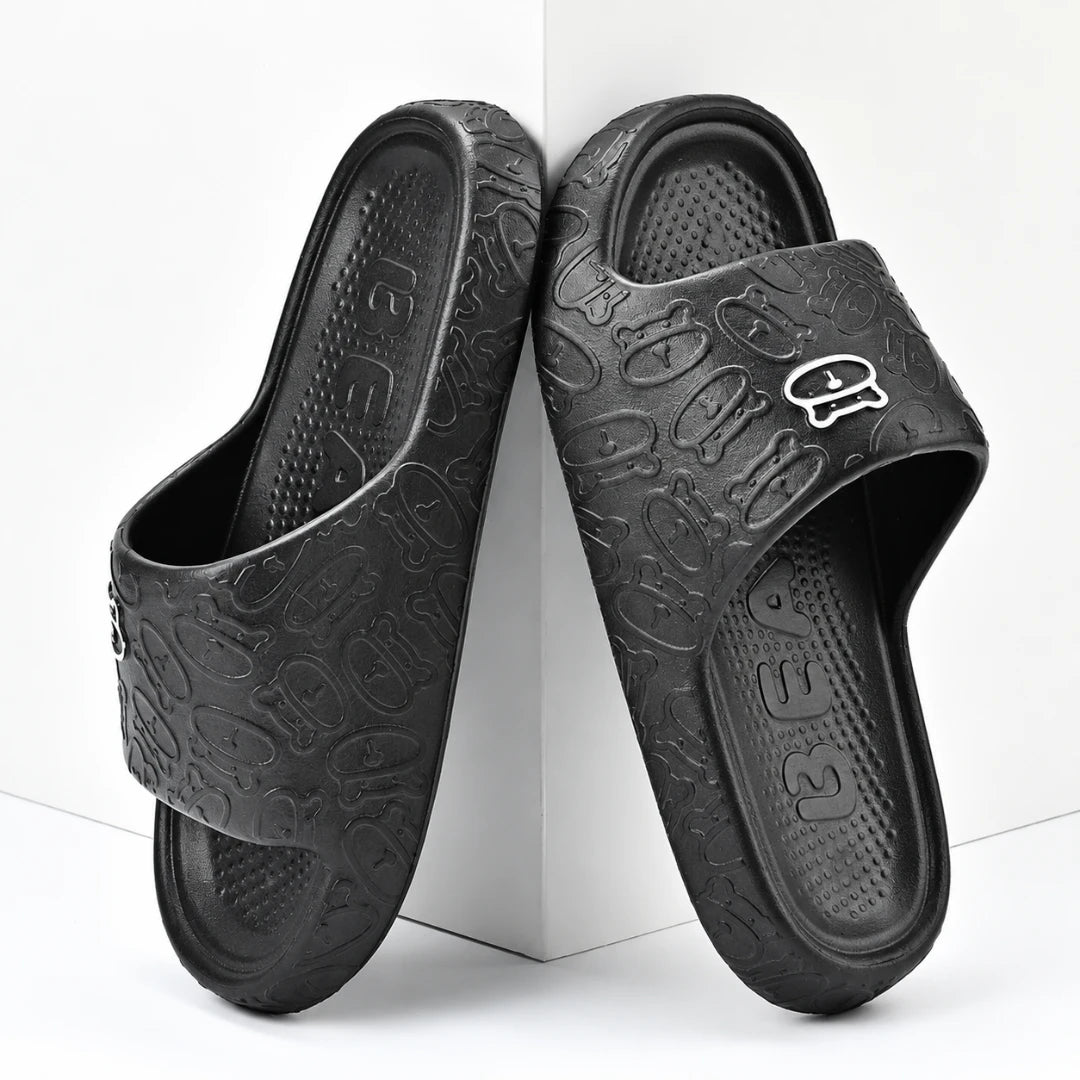 Josh & Tate - SoftBear™ Non-Slip Cushion Slides - A pair of black slide sandals with an embossed cartoon pattern and a small silver logo on the strap, displayed on a white geometric background.