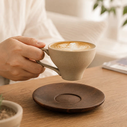Josh & Tate - SoftPour™ Retro Ceramic Latte Cup - A person holds a beige ceramic coffee cup with latte art above a dark brown wooden saucer on a wooden table in a bright indoor setting.