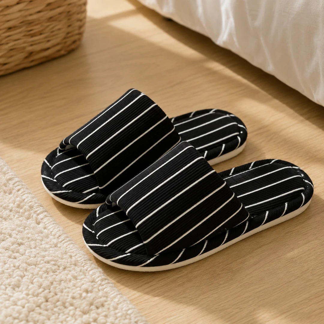 Josh & Tate - SoftStripe™ Cozy Home Slippers - Black slide slippers with thin white vertical stripes, placed on a light wood floor next to a white textured rug and a woven basket.