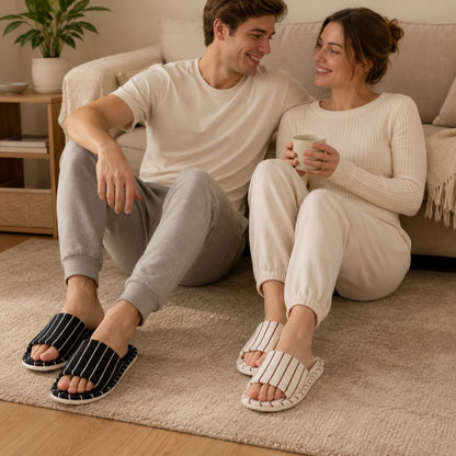 Josh & Tate - SoftStripe™ Cozy Home Slippers - A man and a woman sitting on a beige sofa in a cozy living room, wearing matching cream-colored ribbed long-sleeve tops and jogger pants. The man wears black slip-on slippers with white stripes, and the woman wears white slip-on slippers with thin black stripes.