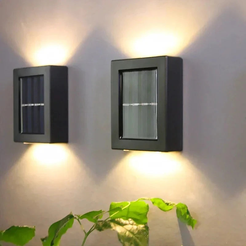 Josh & Tate - SolarGlow Wireless Wall Lamp - Two square black solar wall lights on a gray wall with a plant in the foreground.