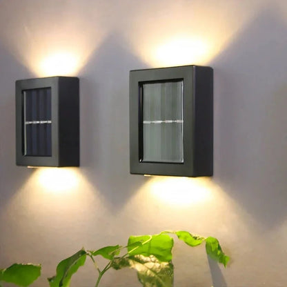 Josh & Tate - SolarGlow Wireless Wall Lamp - Two square black solar wall lights on a gray wall with a plant in the foreground.
