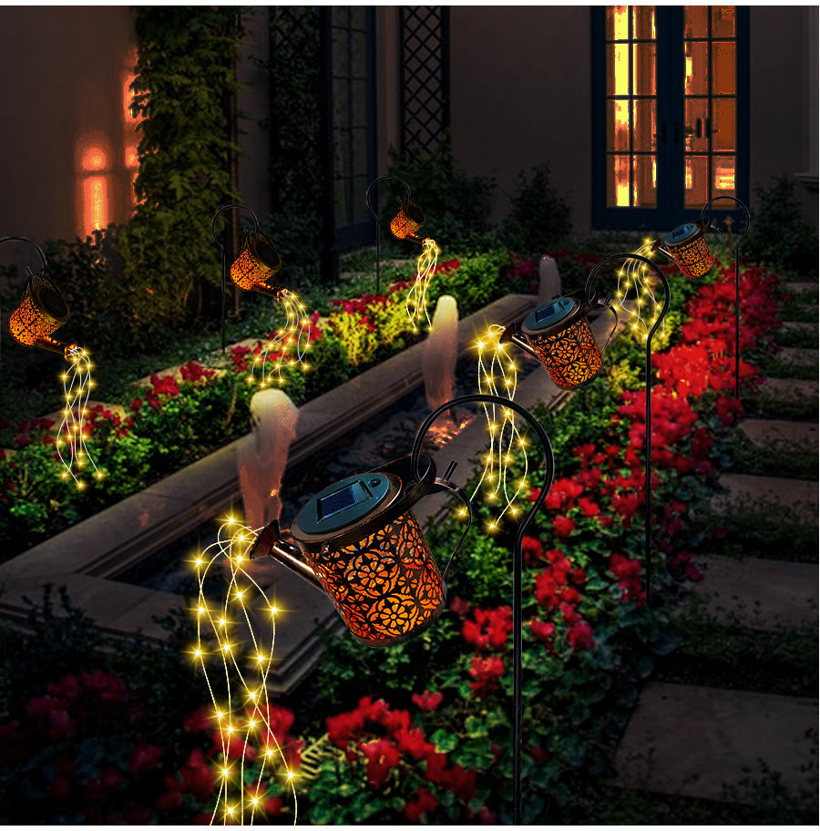Decorative string lights and lanterns on a garden path with flowers and a building in the background.