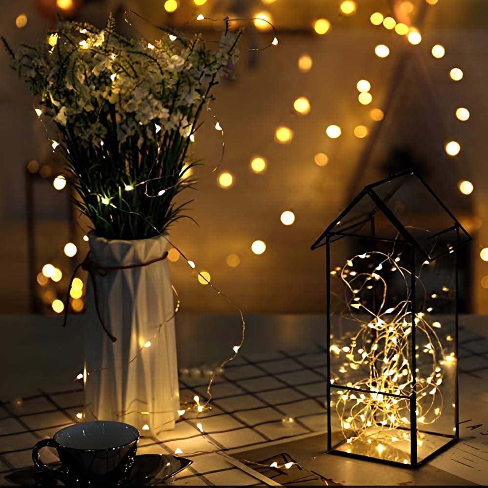 Decorative setup with a vase of flowers, lit lantern, and cup on a table with string lights in the background.