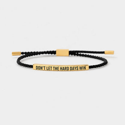 Josh & Tate - StainlessSteel™ Adjustable Bracelet - A black braided cord bracelet with a polished gold-plated bar engraved with the motivational quote 'DON'T LET THE HARD DAYS WIN'. The bracelet is displayed on a clean, light gray background.