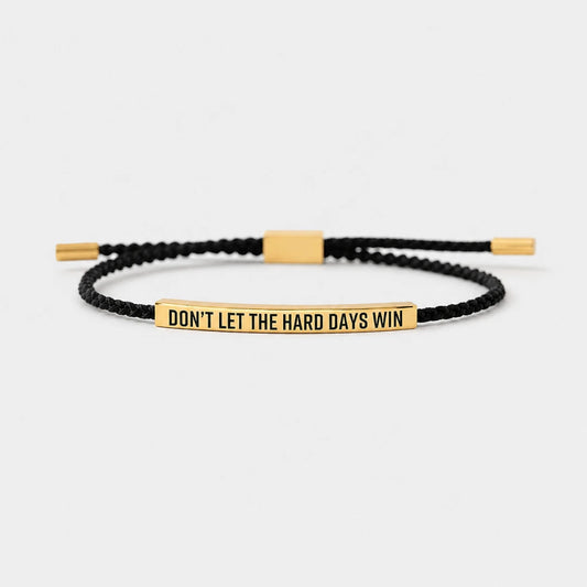 Josh & Tate - StainlessSteel™ Adjustable Bracelet - A black braided cord bracelet with a polished gold-plated bar engraved with the motivational quote 'DON'T LET THE HARD DAYS WIN'. The bracelet is displayed on a clean, light gray background.