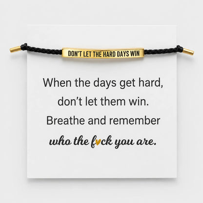 Josh & Tate - StainlessSteel™ Adjustable Bracelet - A black cord bracelet with a gold-toned rectangular bar engraved with the phrase 'DON'T LET THE HARD DAYS WIN' is displayed across a white card. The card also features a motivational quote printed in black text, with the word 'fuck' highlighted in gold.