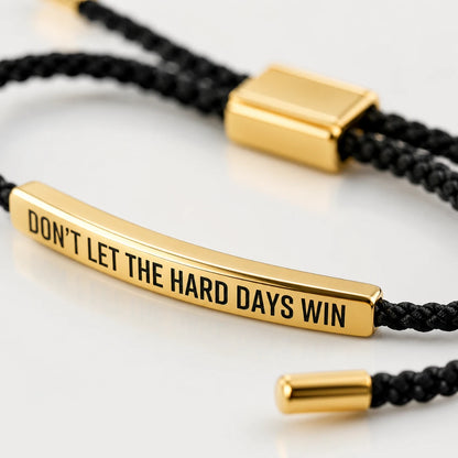 Josh & Tate - StainlessSteel™ Adjustable Bracelet - A close-up of a black braided cord bracelet with a polished gold bar engraved with the motivational quote 'DON'T LET THE HARD DAYS WIN'.
