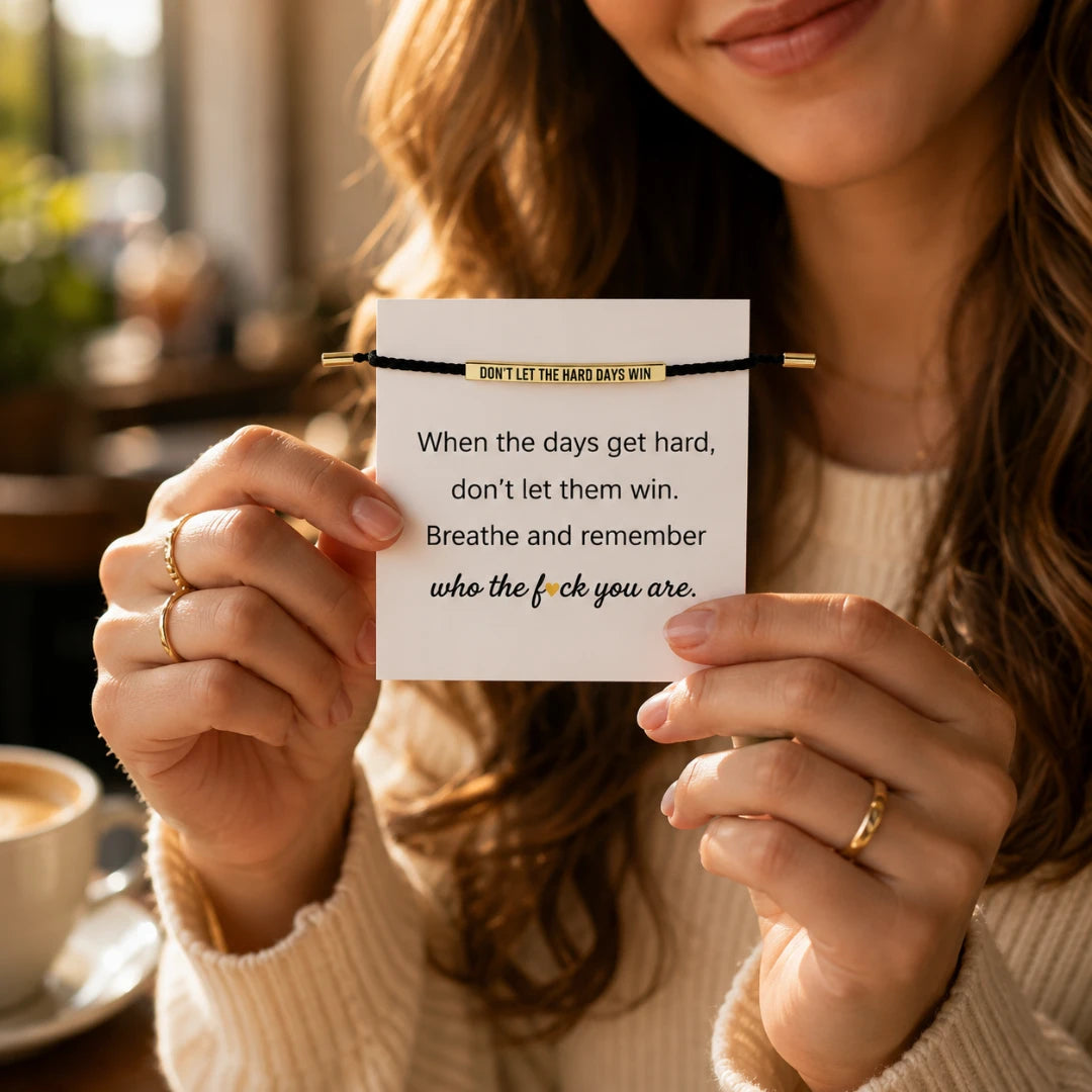 Josh & Tate - StainlessSteel™ Adjustable Bracelet - A woman in a cream knit sweater holds up a black cord bracelet with gold-toned ends and a gold banner that reads 'DON'T LET THE HARD DAYS WIN' in black text. The background is a warm, softly blurred indoor setting with a coffee cup visible on the left.