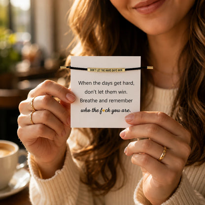 Josh & Tate - StainlessSteel™ Adjustable Bracelet - A woman in a cream knit sweater holds up a black cord bracelet with gold-toned ends and a gold banner that reads 'DON'T LET THE HARD DAYS WIN' in black text. The background is a warm, softly blurred indoor setting with a coffee cup visible on the left.
