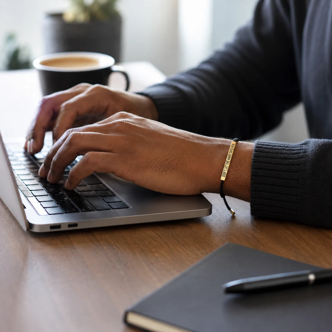 Josh & Tate - StainlessSteel™ Adjustable Bracelet - A close-up lifestyle shot of a person typing on a laptop at a wooden desk, wearing a gold bar bracelet with engraved text on their wrist.