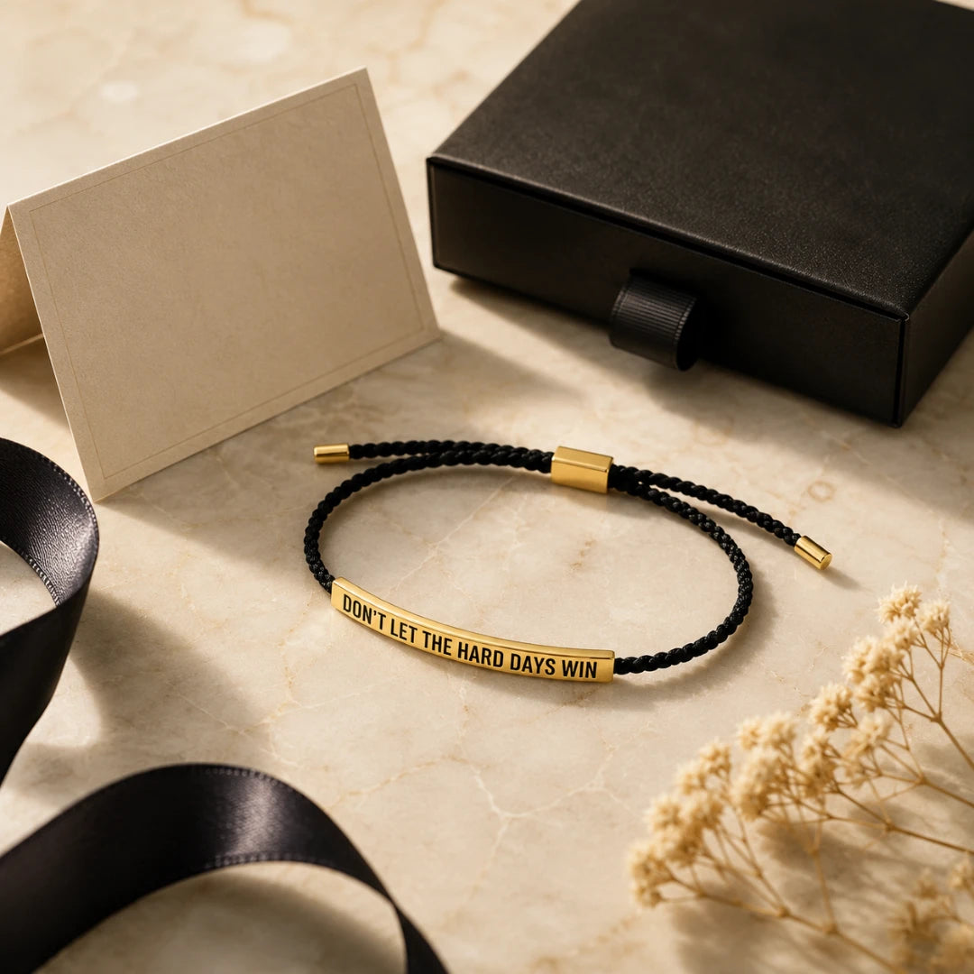 Josh & Tate - StainlessSteel™ Adjustable Bracelet - A black braided cord bracelet with a gold bar engraved with the phrase 'DON'T LET THE HARD DAYS WIN', styled on a light marble surface with a black gift box, ribbon, and dried flowers.
