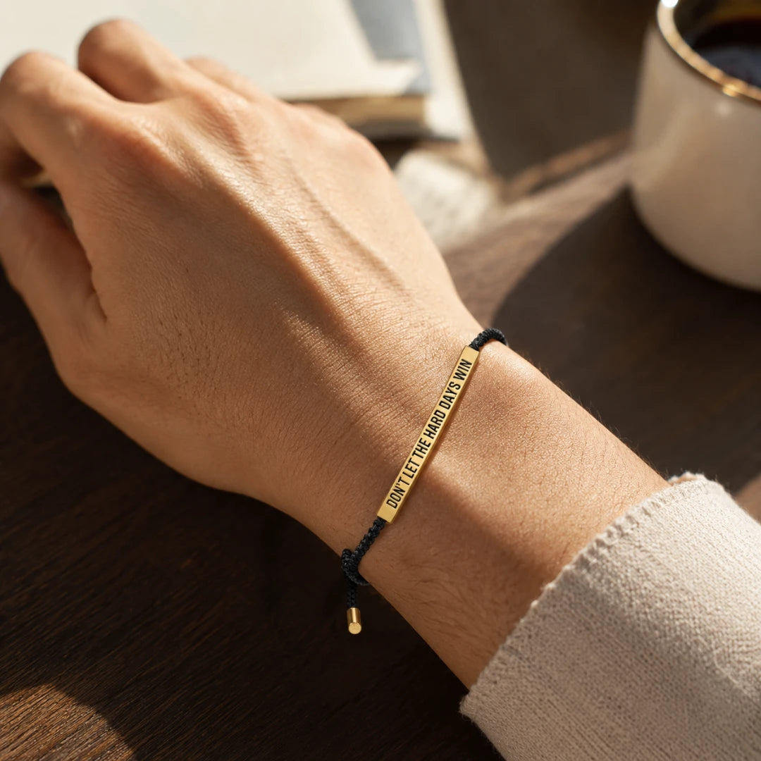 Josh & Tate - StainlessSteel™ Adjustable Bracelet - A close-up of a person's wrist wearing a gold-plated engraved bracelet with a black cord, resting on a dark wooden table next to a white mug.