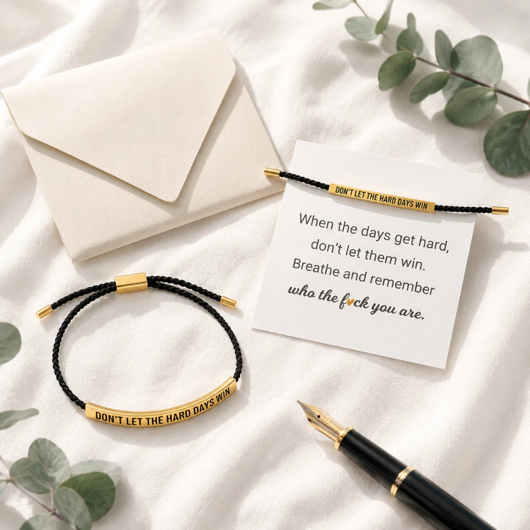 Josh & Tate - StainlessSteel™ Adjustable Bracelet - Two black braided bracelet-style necklaces with gold-toned rectangular bars engraved with the phrase 'DON'T LET THE HARD DAYS WIN' are arranged on a white fabric surface with a cream envelope, a printed quote card, green leaves, and a black-and-gold pen.