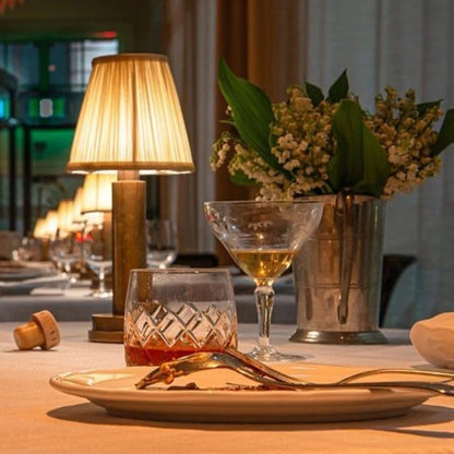 Dining table setting with glasses, a vase of flowers, and a lamp in a warm indoor setting.