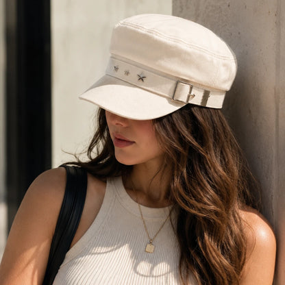 Josh & Tate - StarHat™ Retro Beret - A woman wearing a cream-colored newsboy cap with a silver buckle and star details, leaning against a concrete wall.
