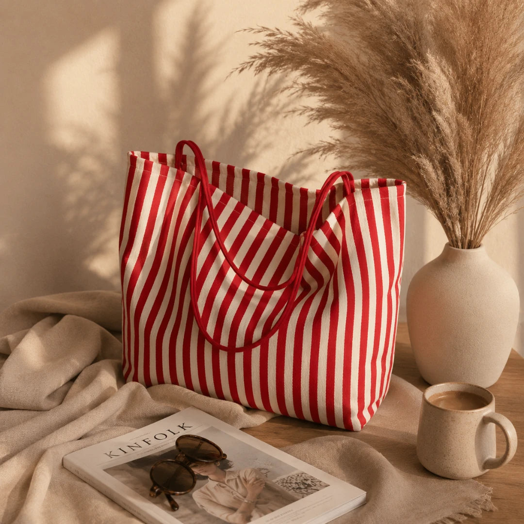 A red and white vertically striped canvas tote bag with red handles, styled on a wooden table with a beige throw blanket, sunglasses on a magazine, a ceramic mug, and a vase of pampas grass against a sunlit beige wall.