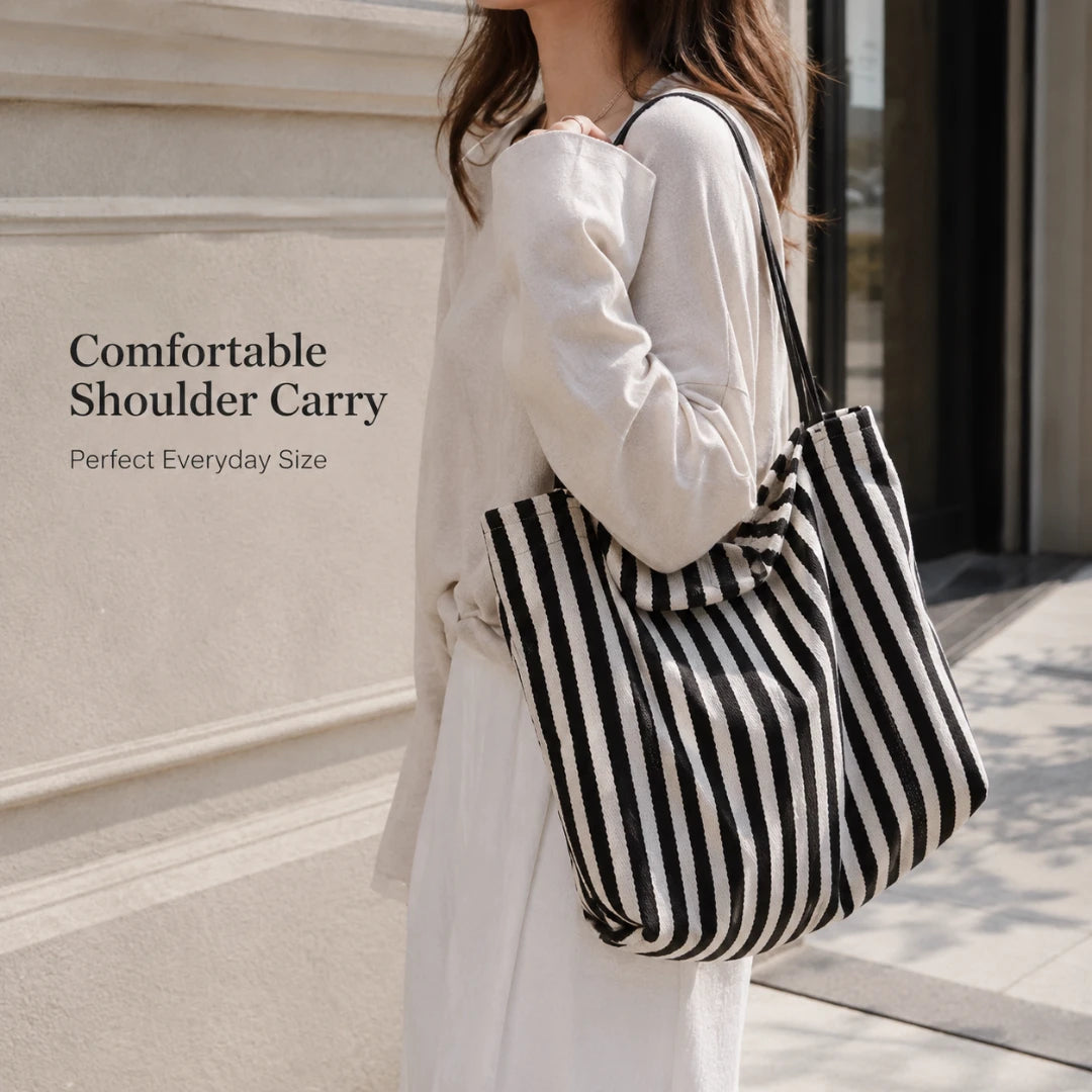 Josh & Tate - StripeCarry™ Oversized Tote Bag - A woman in a white dress carries a large black and white striped shoulder tote bag outdoors against a light stone wall. Text on the left reads 'Comfortable Shoulder Carry' and 'Perfect Everyday Size'.
