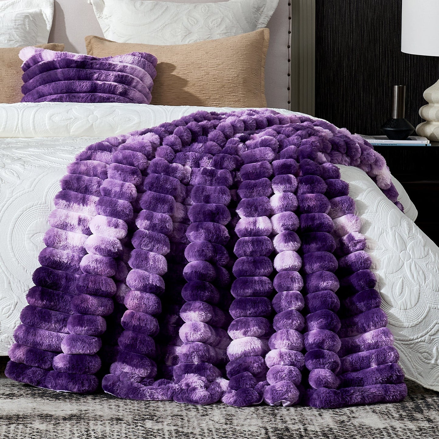 Purple fuzzy blanket draped over a bed with pillows and a lamp in the background.