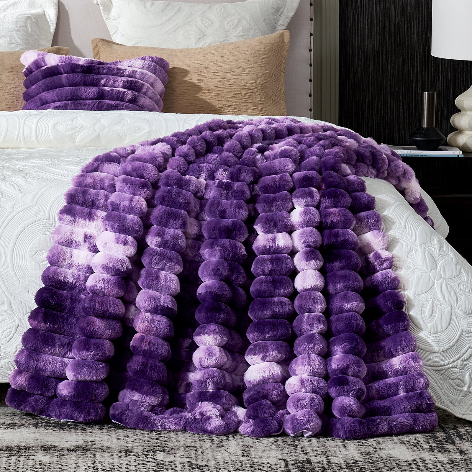Purple fuzzy blanket draped over a bed with pillows and a lamp in the background.