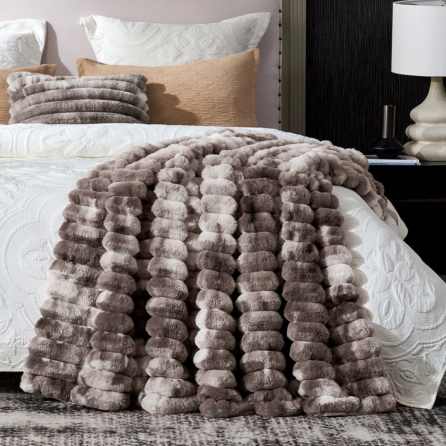 Fluffy gray blanket draped over a bed with pillows and a lamp in the background.