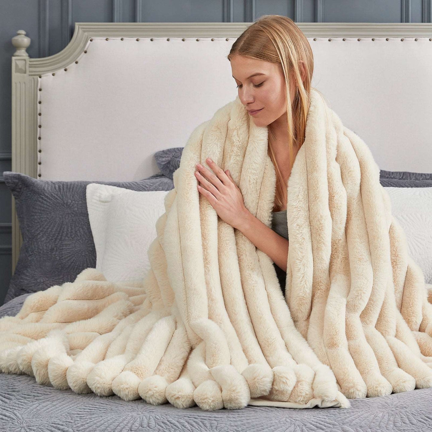 Woman wrapped in a plush beige blanket on a bed with a neutral headboard.