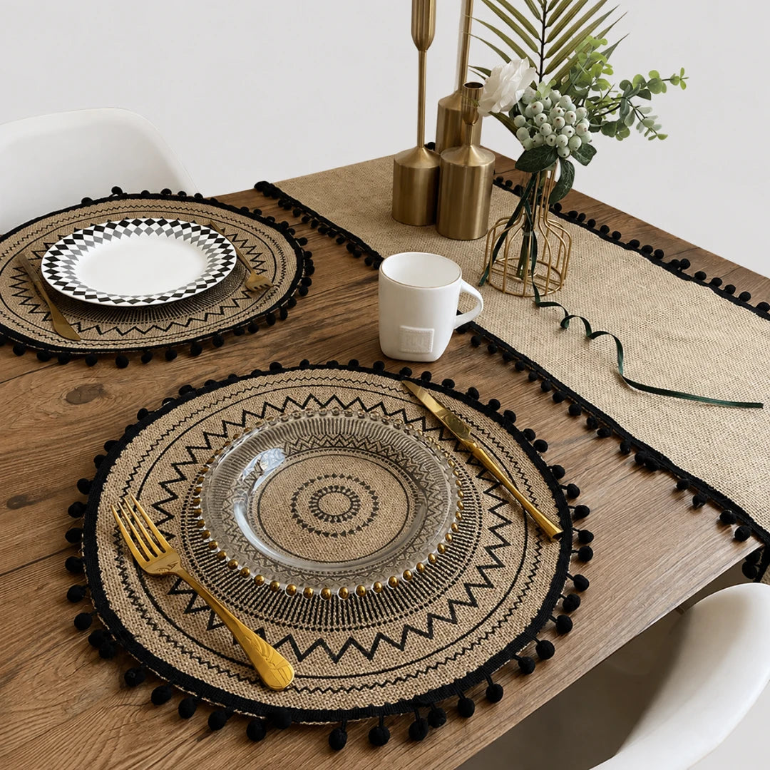 Josh & Tate - SunWeave™ Boho Mandala Placemat - A styled dining table setting featuring round woven placemats with black pom-pom trim, a matching table runner, gold-toned cutlery, and clear glass plates on a wooden table.